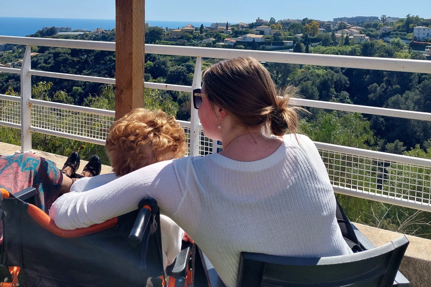 Junge Frau legt Arm um ältere Person im Rollstuhl auf Terrasse mit Meer- und Hügelblick bei Nizza.