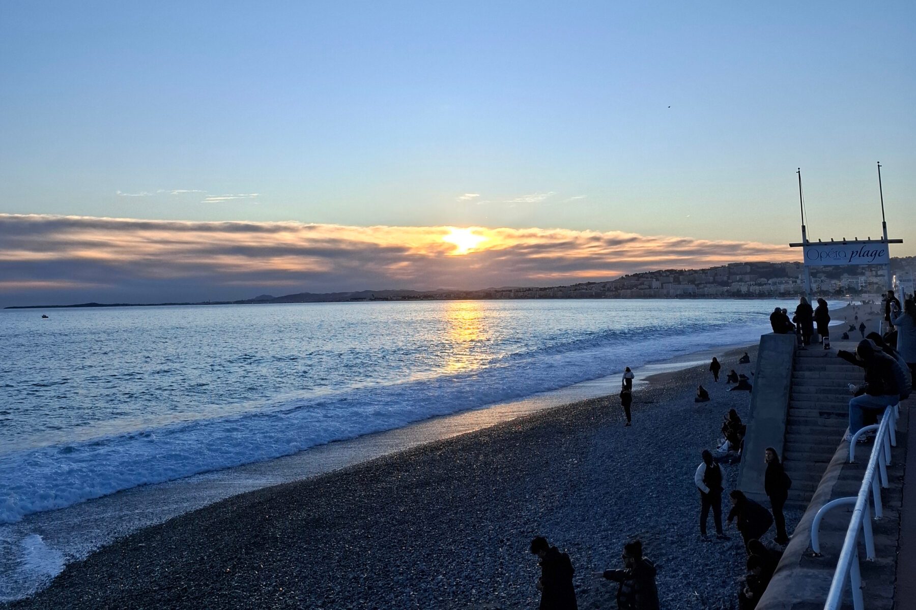 Sonnenuntergang über dem Meer bei La Colline in Nizza, Kiesstrand mit Wellen und Silhouetten von Menschen auf Promenade