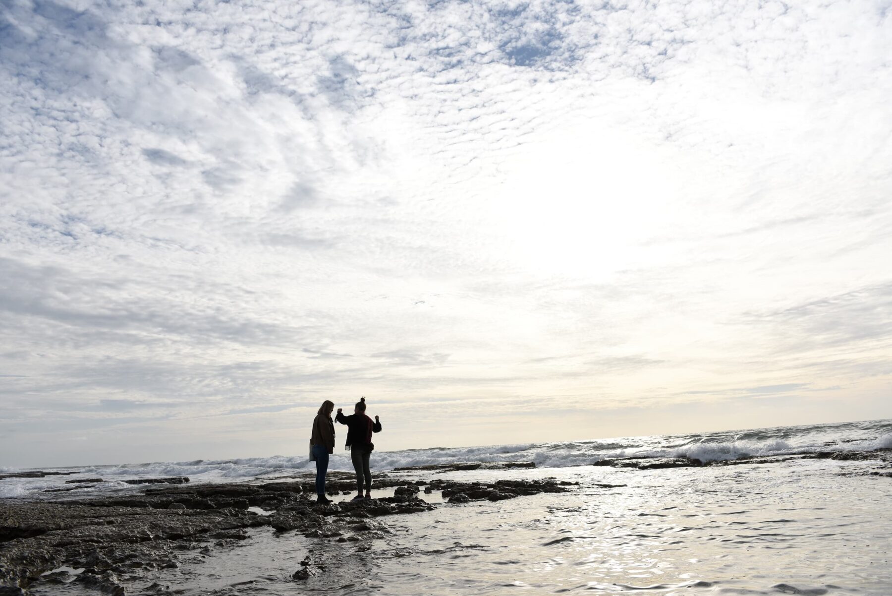 Zwei Personen stehen auf felsigem Ufer, Silhouetten vor weitem bedecktem Himmel und glänzendem Meer.