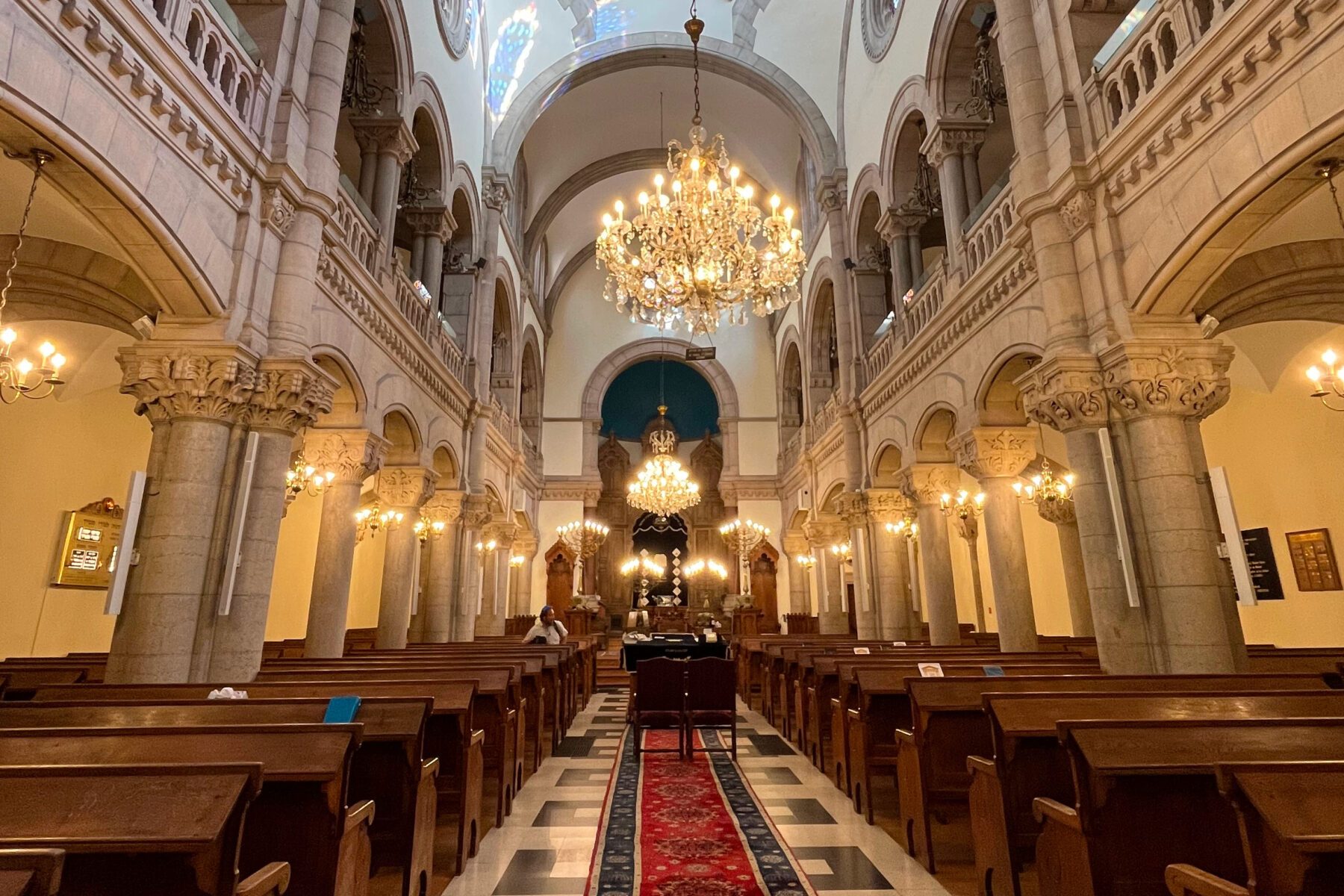 Innenraum der Großen Synagoge von Lyon mit mittigem roten Teppich, Holzbankreihen, Säulen und opulenten Kronleuchtern.