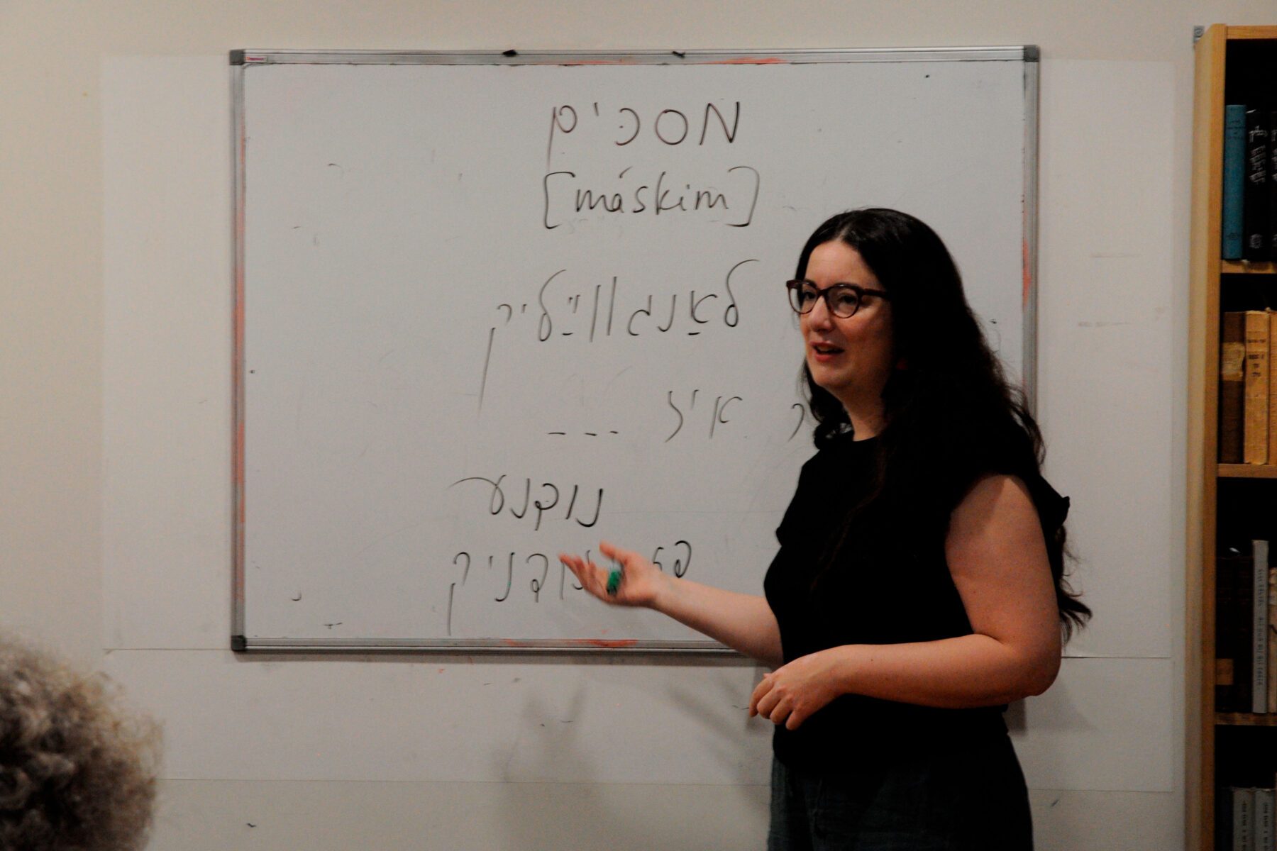 Lehrerin zeigt mit Marker jiddische Schrift auf Whiteboard in Pariser Klassenzimmer, Bücherregal rechts
