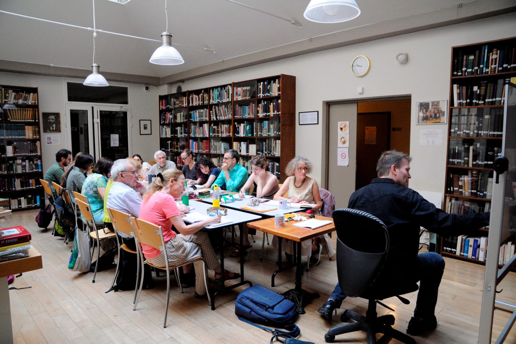 Erwachsene in Bibliothek sitzen an langen Tischen beim Unterricht, Bücherregale an den Wänden, Lehrende rechts am Whiteboard