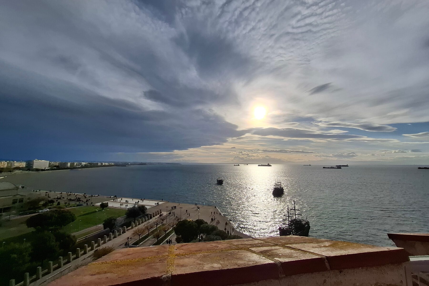 Sonnenreflexe auf dem Meer bei Thessaloniki, dramatische Wolken, Schiffe und Menschen auf der Uferpromenade.