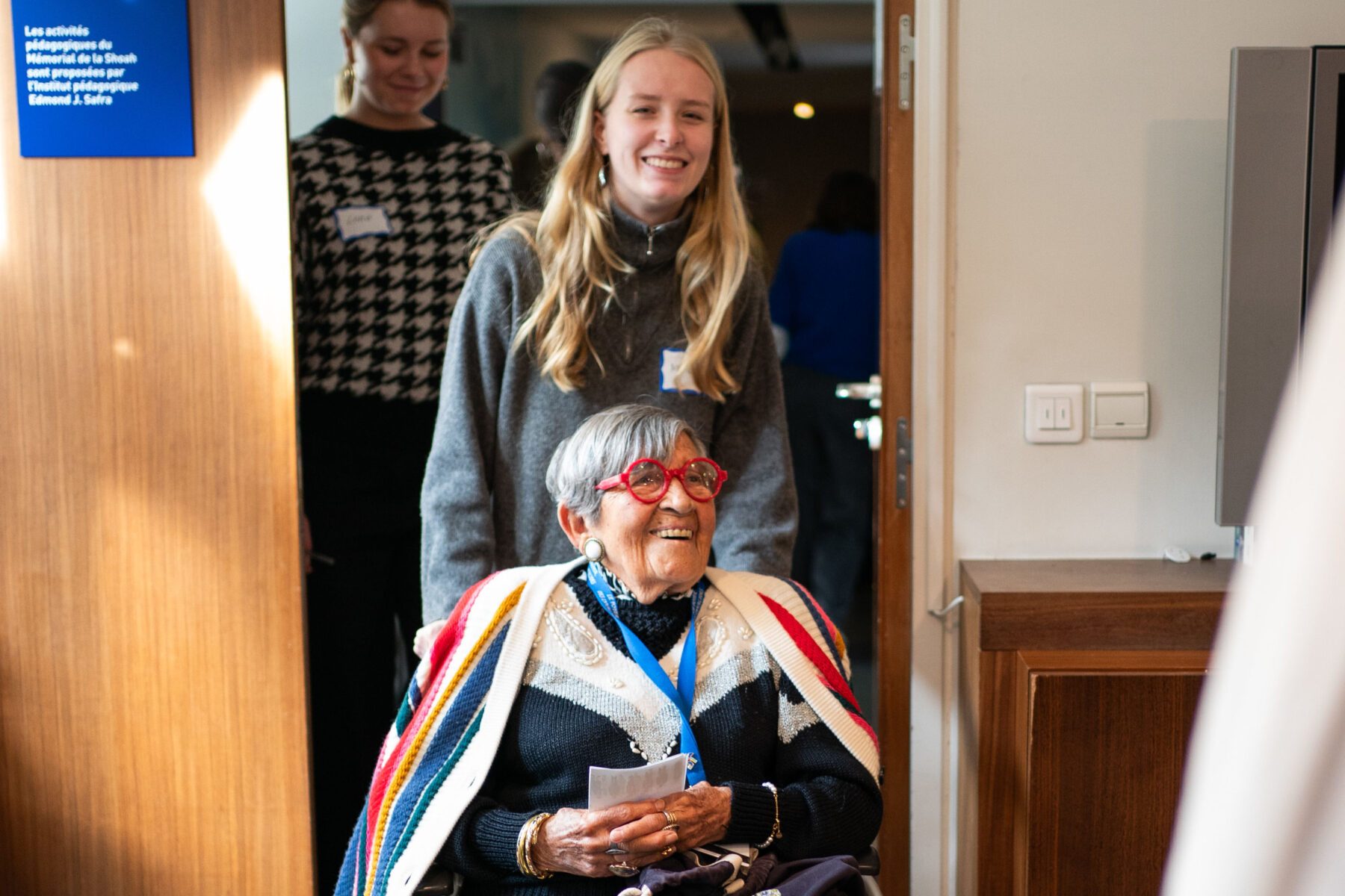 Ältere Frau im Rollstuhl mit buntem Schal und Namensbadge, hält Zettel; junge Begleiterin dahinter im Mémorial de la Shoah