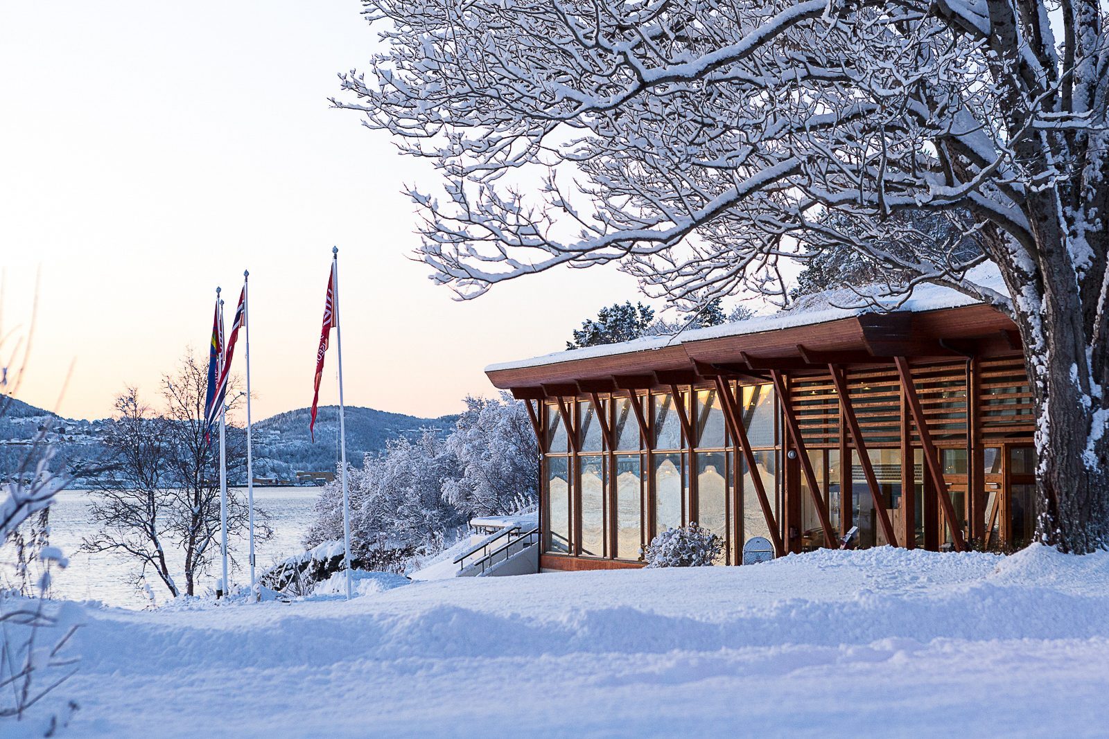 Trondenes Museum am verschneiten Ufer, modernes Holzgebäude mit großen Fenstern, Flaggenmasten und schneebedeckte Bäume.