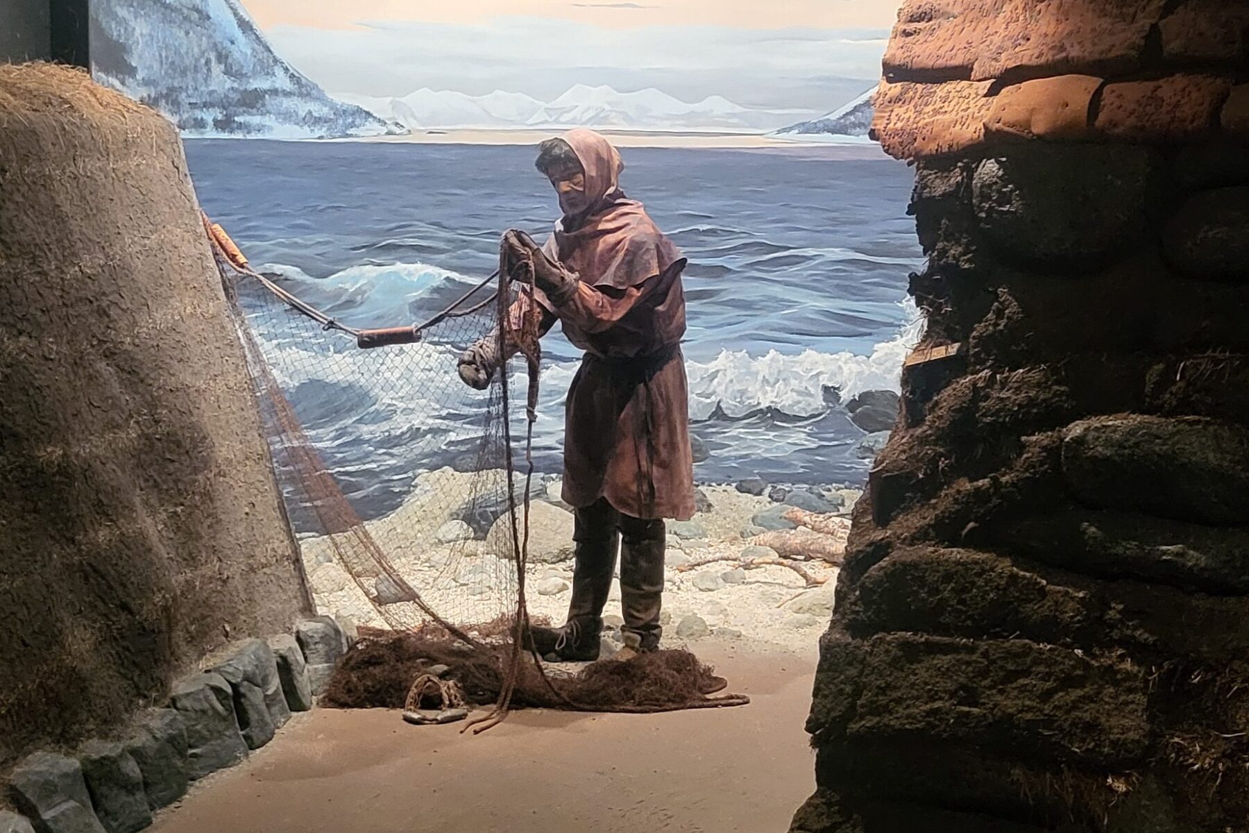 Ausstellung im ASF-Museum Trondenes: Fischerfigur in brauner Kapuzenjacke flickt Netz vor felsiger Küste und Wellen.