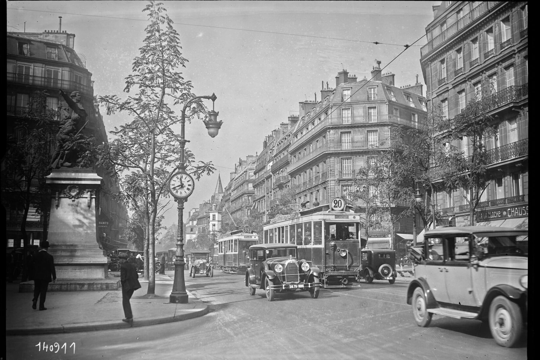 Boulevard Saint‑Germain 1929: Straßenbahn und Oldtimer vor Haussmann‑Häusern, Standuhr, Denkmal und Fußgänger.