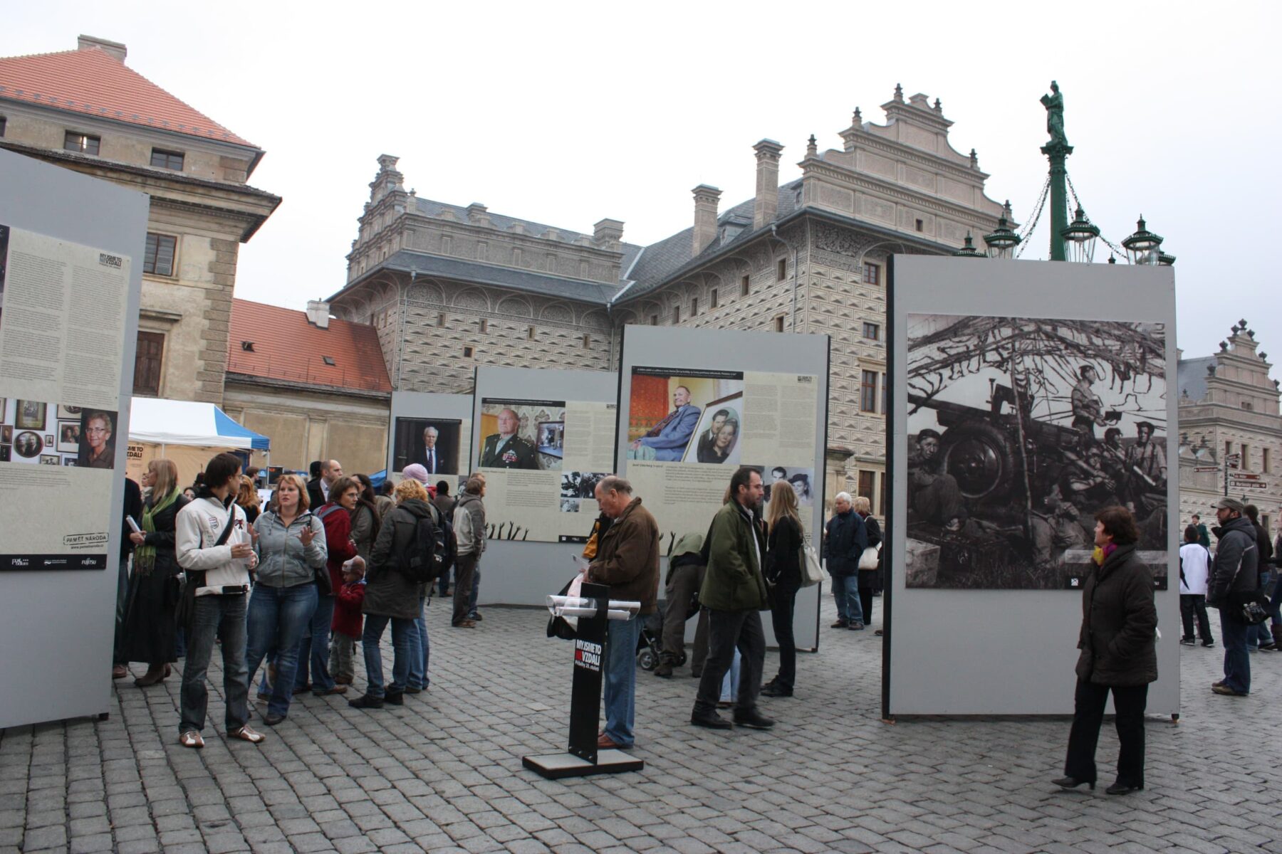 Freiluft-Fotoausstellung 'post bellum' auf gepflastertem Platz vor Renaissancegebäude, Besucher betrachten Infotafeln.