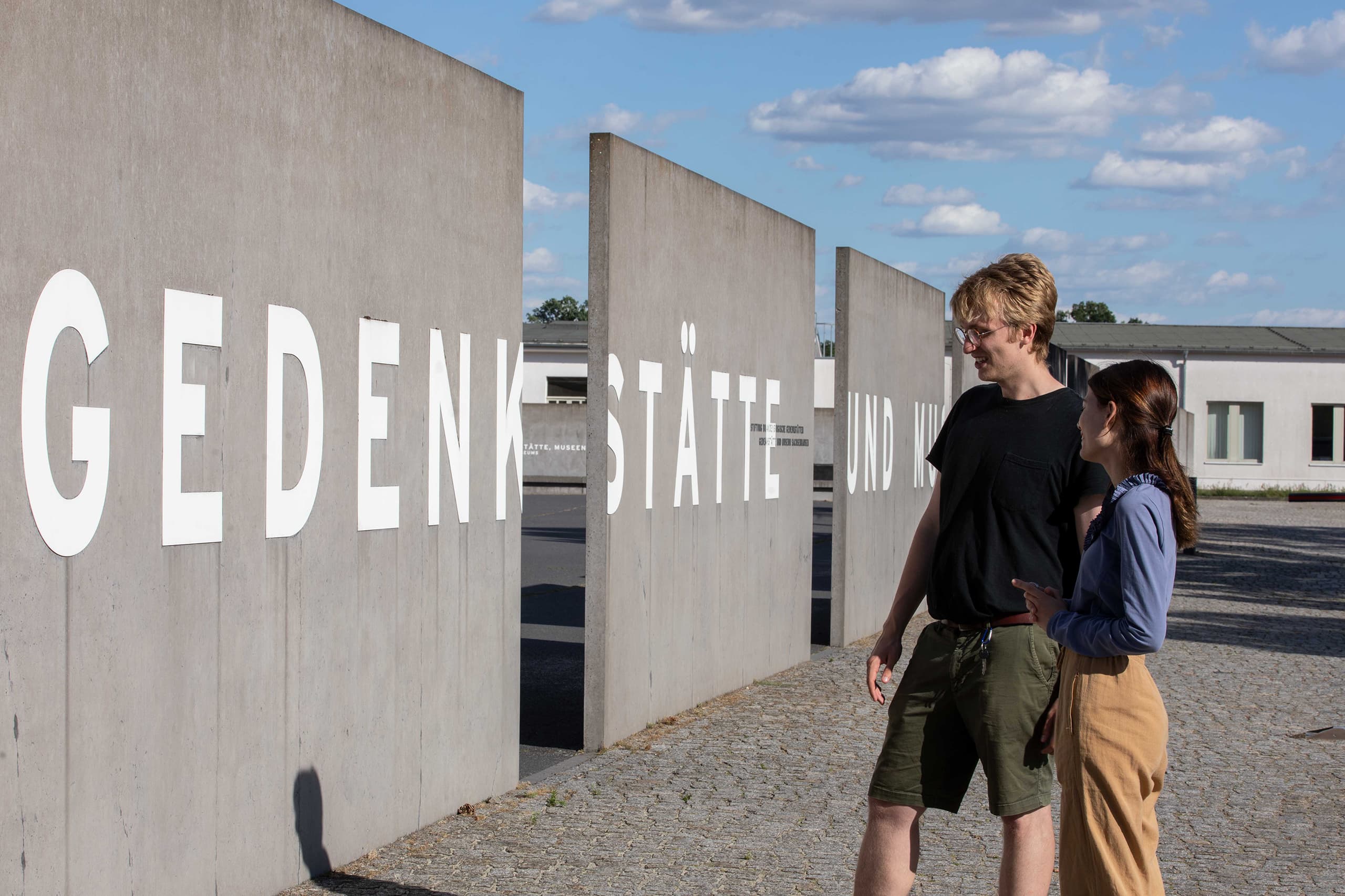 Zwei Menschen besuchen die Gedenkstätte Sachsenhausen in Deutschland. Große Buchstaben auf der Betonwand. Sonne scheint.