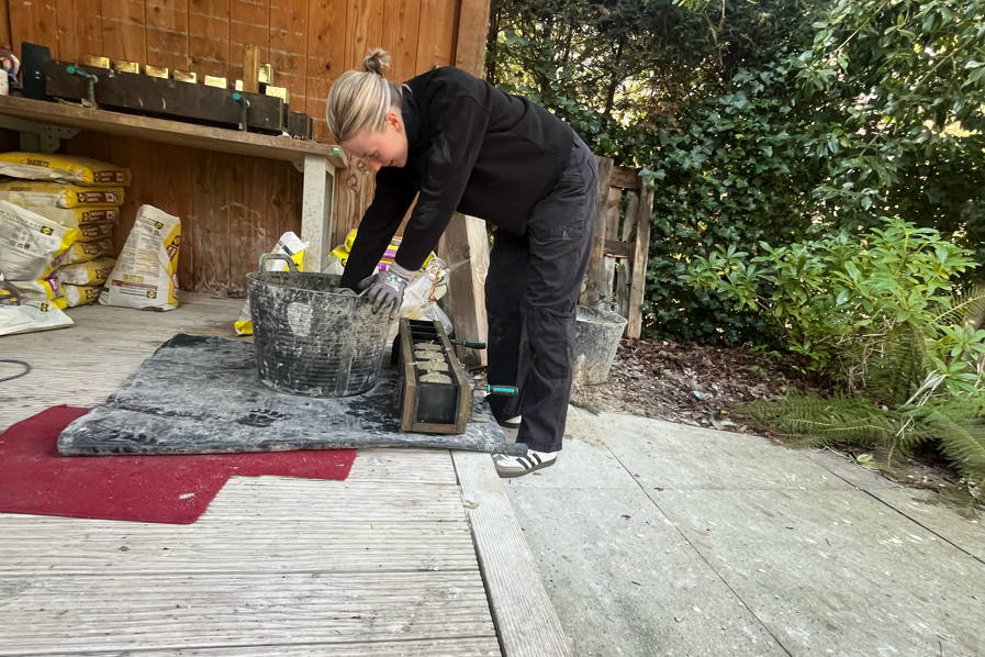 Person in schwarzer Kleidung mit Dutt kniet über einem Eimer und mischt Mörtel neben einer Form für Stolpersteine im Garten