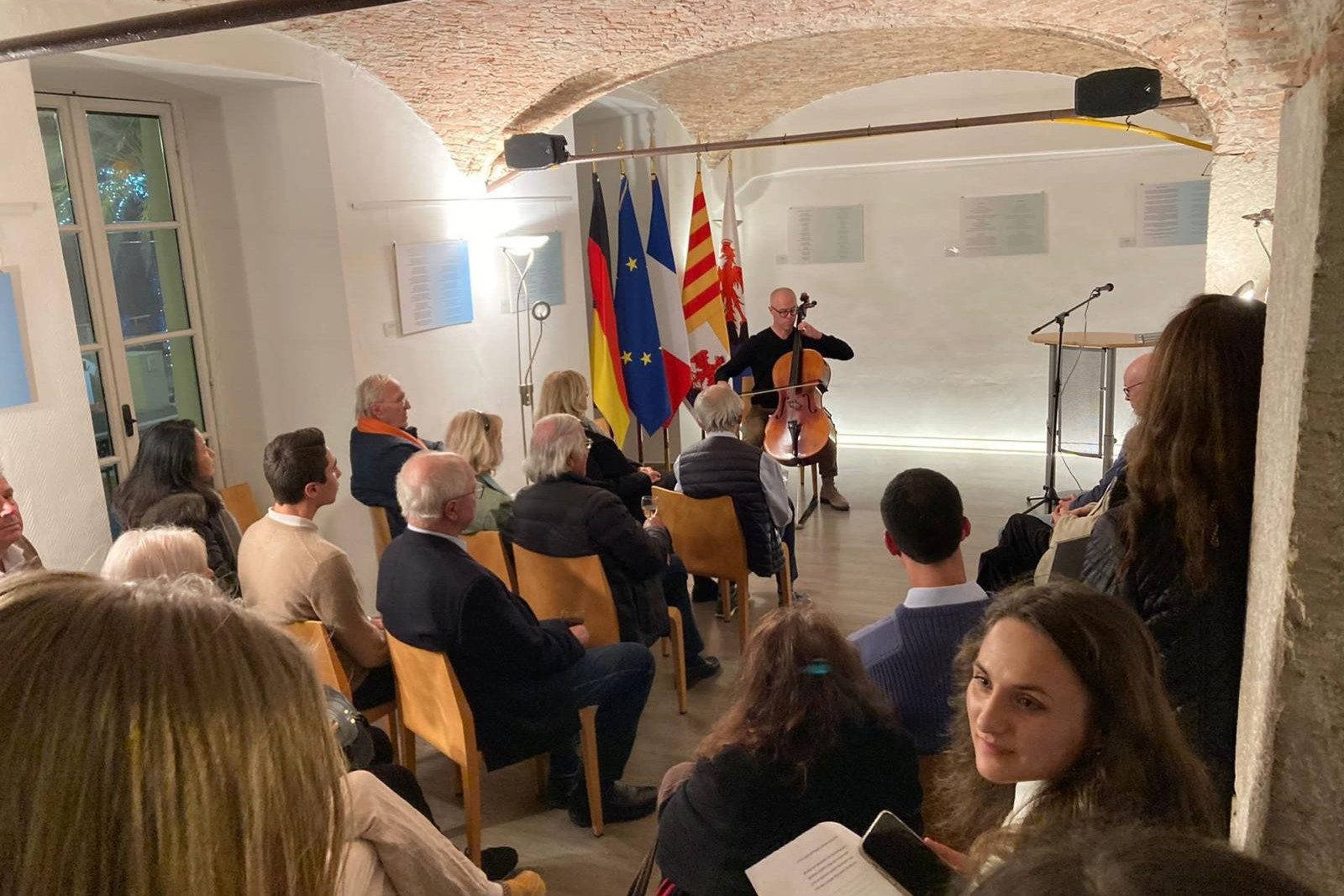 Cellist spielt in kleinem Gewölberaum vor sitzendem Publikum; deutsche, französische und EU-Flaggen im Hintergrund.