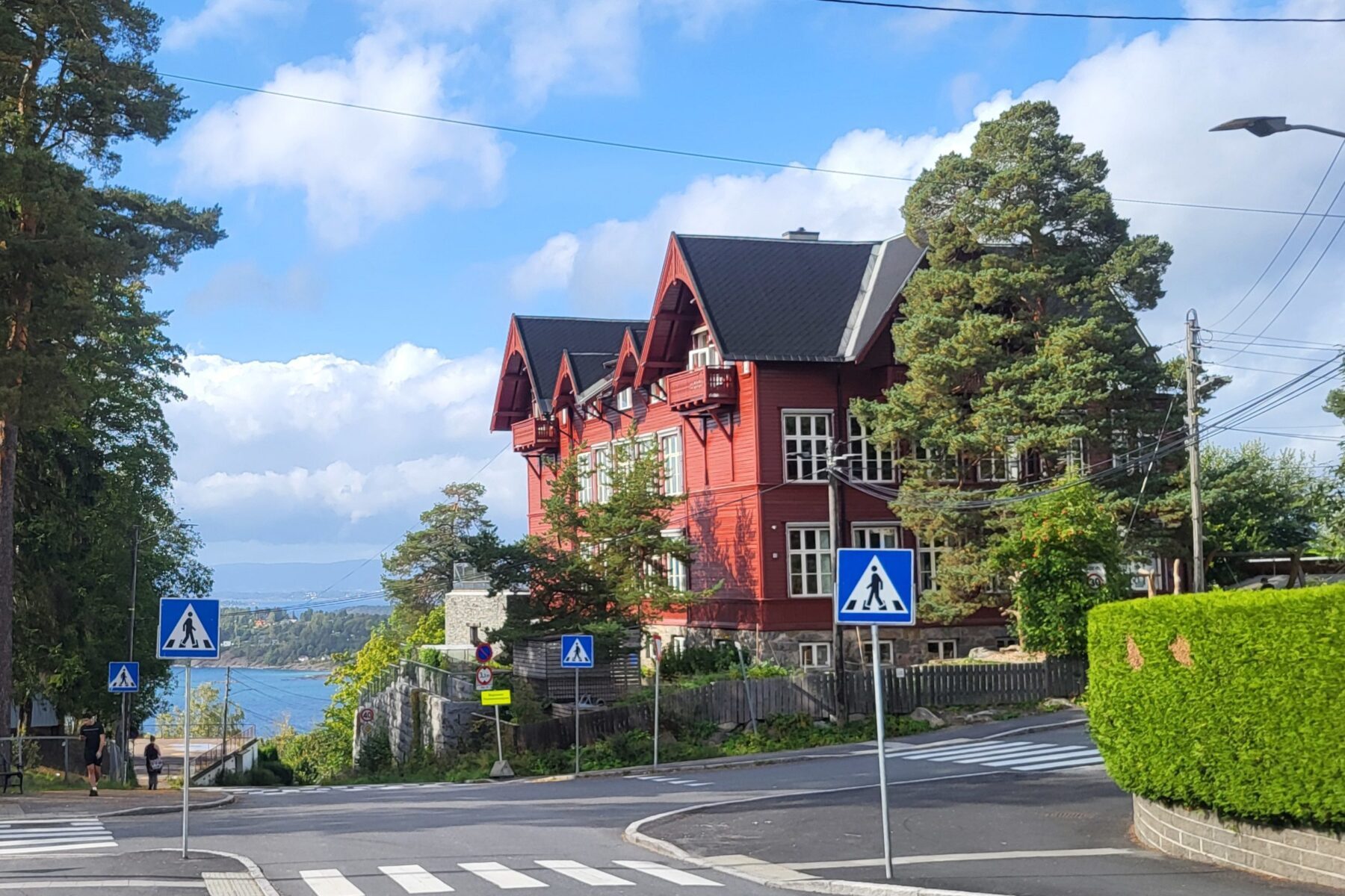 Rotes Holzgebäude (Stiftung Signo) an Hang in Oslo, Zebrastreifen, Fußgänger‑Schilder, Meerblick.