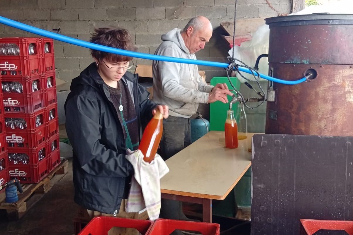 ASF‑Freiwillige im Bocage füllen Glasflaschen mit orangefarbenem Saft an einem Metallbehälter und blauen Schläuchen.