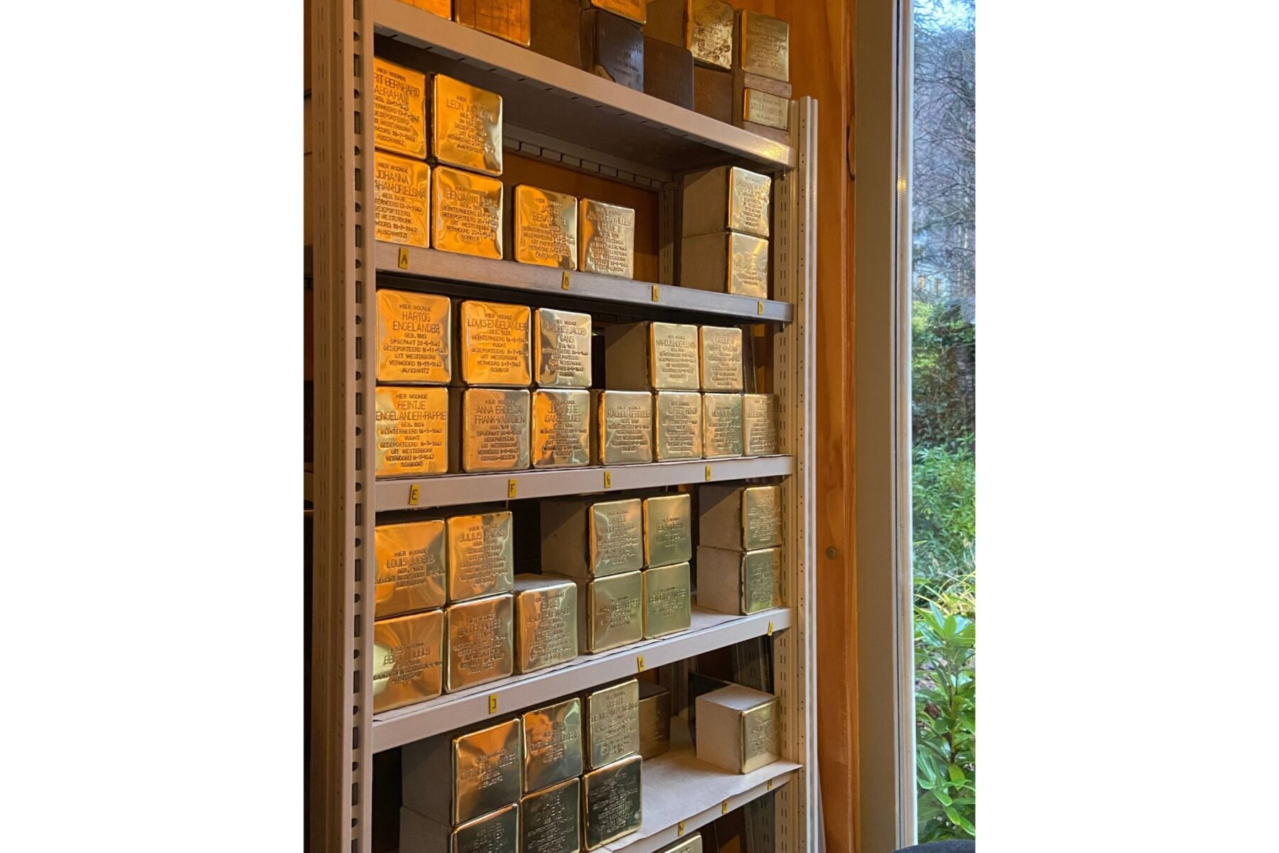 Regal mit gestapelten polierten Messingwürfeln (Stolpersteine) mit eingravierten Namen; rechts Fenster zum Garten.