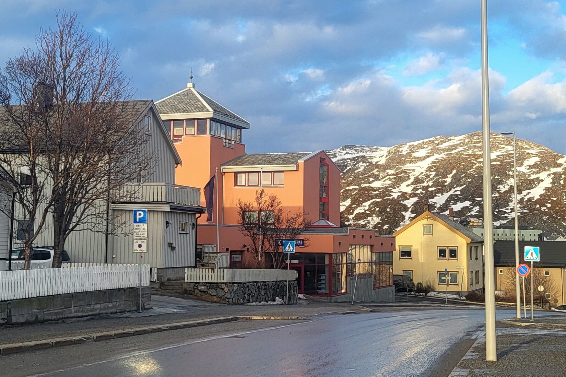 Wiederaufbaumuseum Hammerfest: rosa Gebäude vor schneebedecktem Berg, nasse Straße mit Fußgängerüberweg