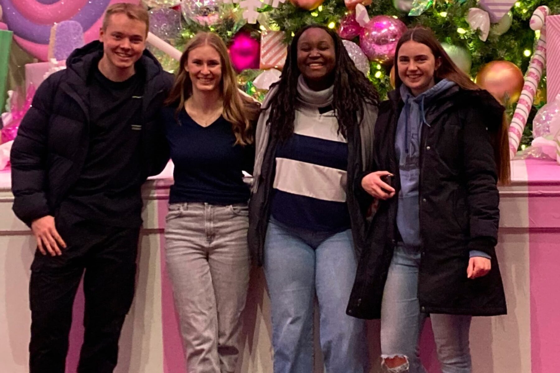 Vier lächelnde Teilnehmende vor einem pink geschmückten Weihnachtsbaum, in Winterjacken und Jeans, festliche Kugeln und Lichter.
