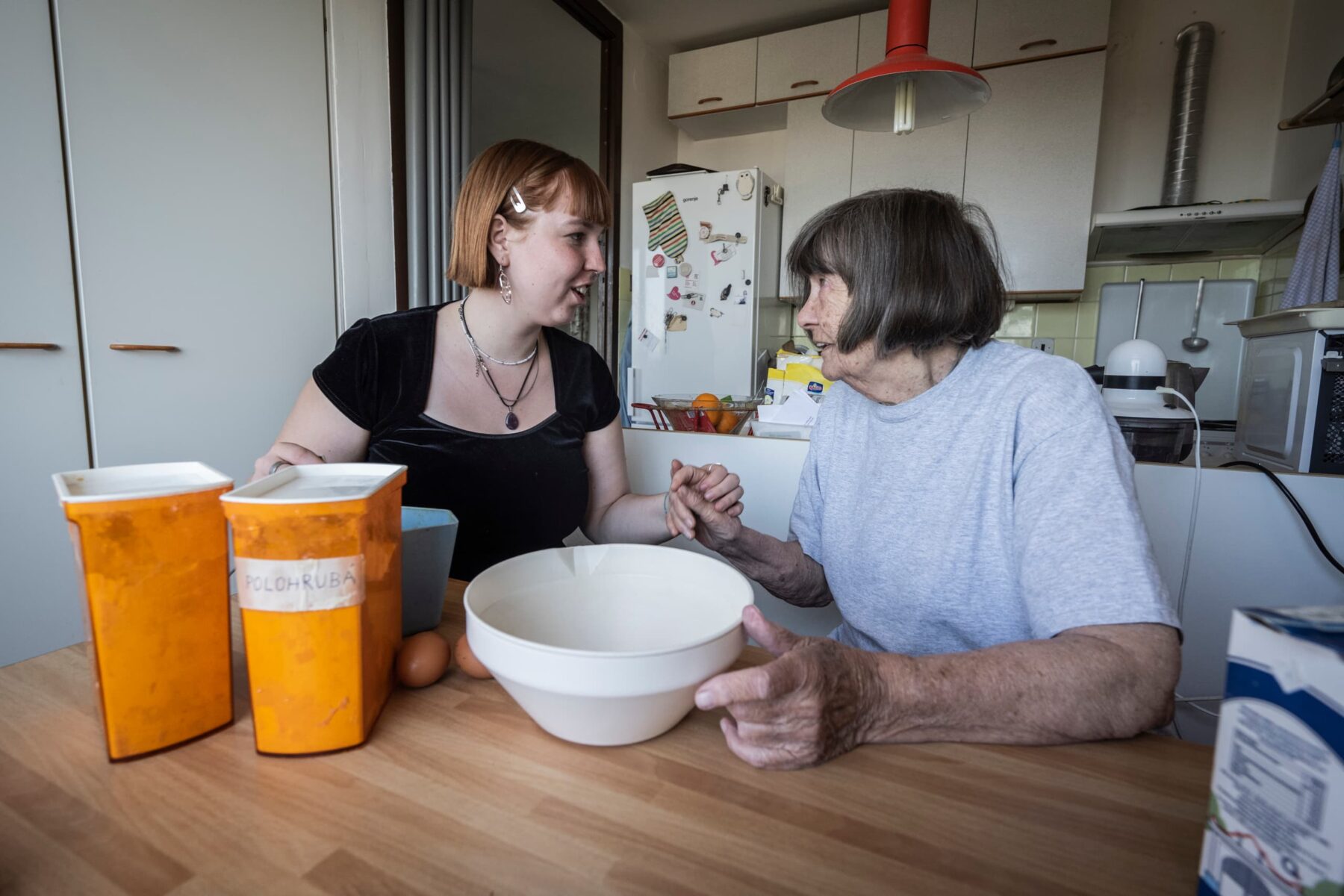 Freiwillige hält in Prager Küche die Hand einer älteren Frau; Schüssel, orange Vorratsdosen und Eier auf Tisch.