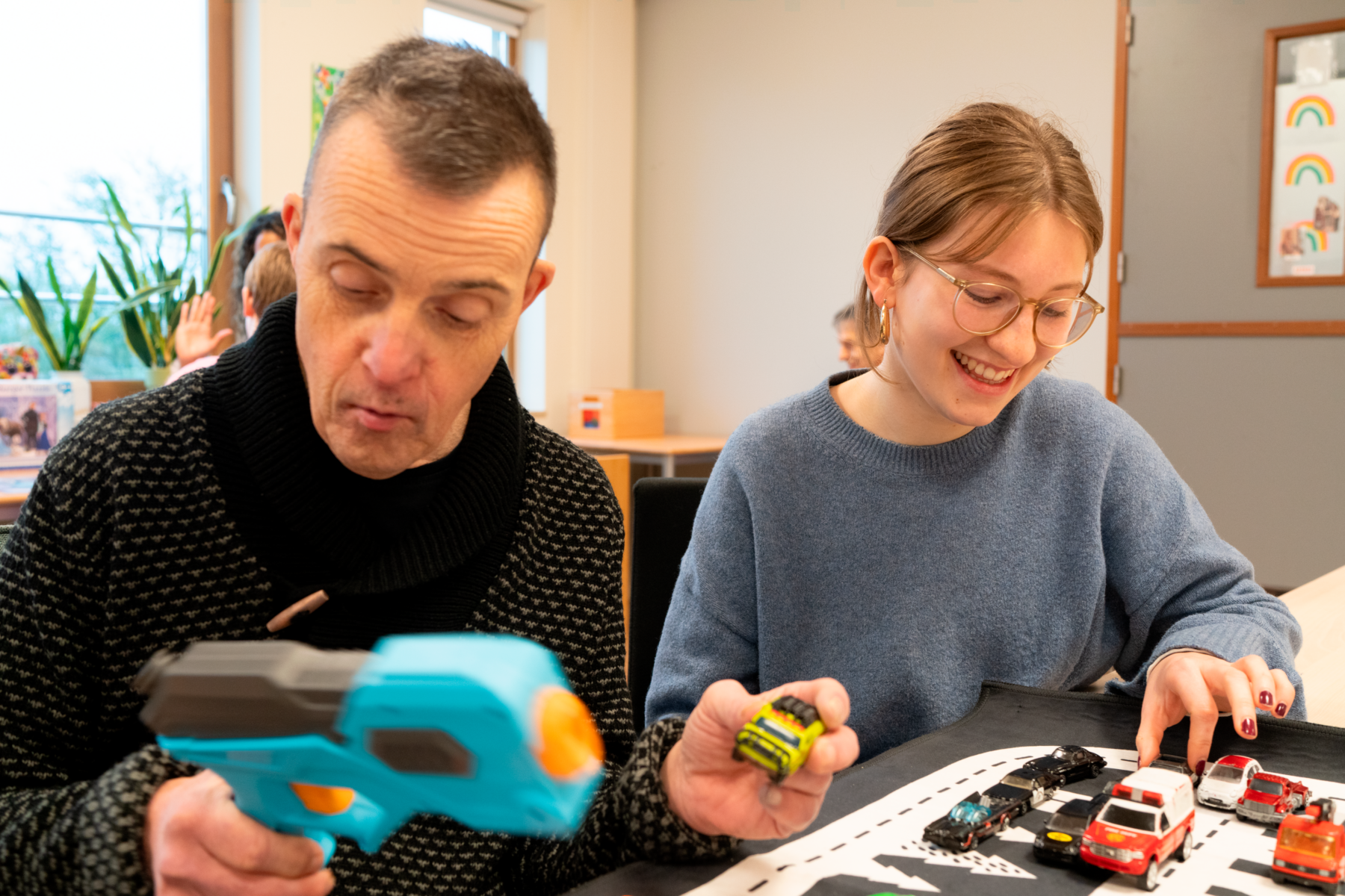 Zwei Freiwillige im Tageszentrum spielen mit Spielzeugautos auf einer Papierstraße; eine hält einen türkis-orangen Blaster.