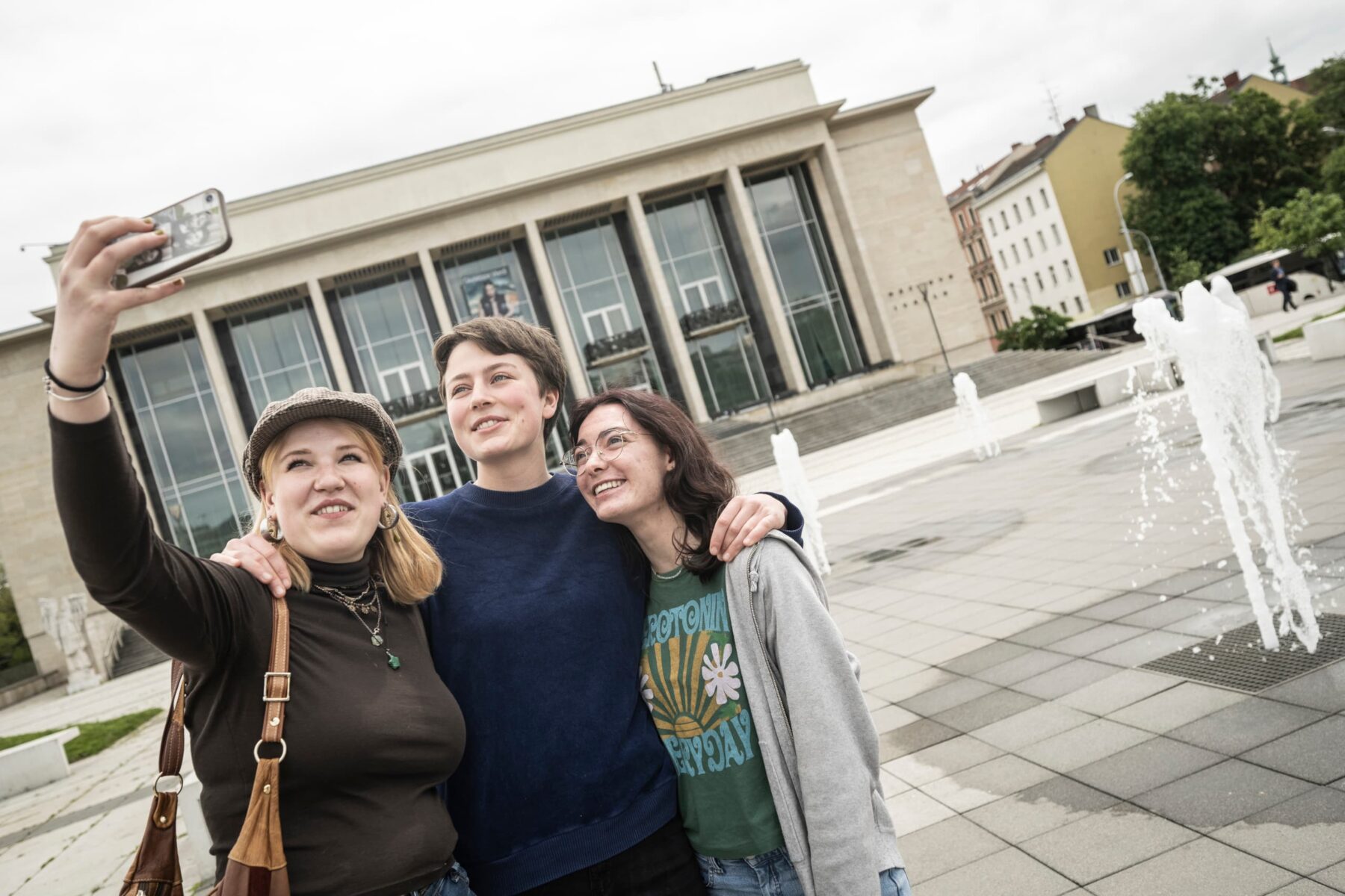 Drei lächelnde ASF-Freiwillige in Brünn machen Selfie vor einem Säulenbau mit Springbrunnen.