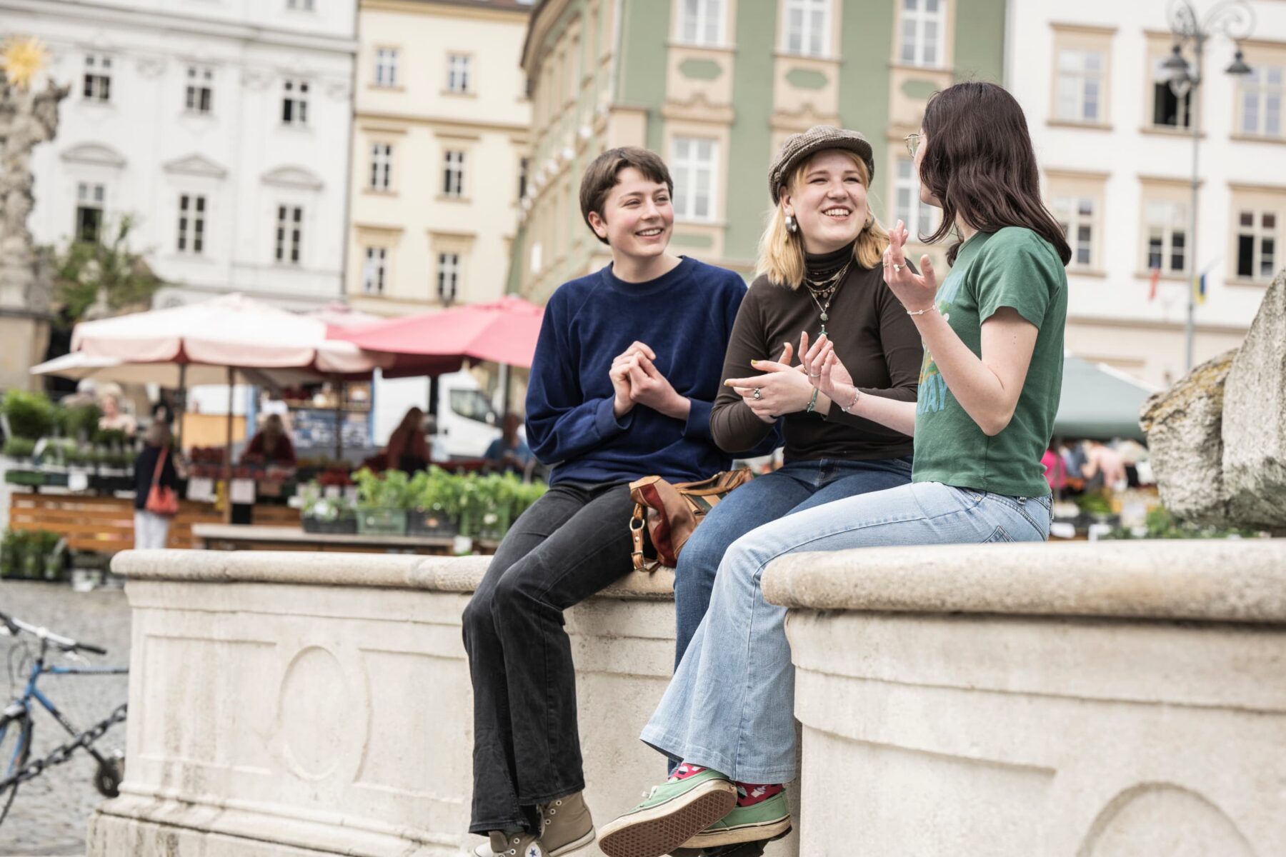 Drei Freiwillige sitzen lachend auf einem Brunnenrand auf dem Marktplatz in Brno, Marktstände und Sonnenschirme im Hintergrund.