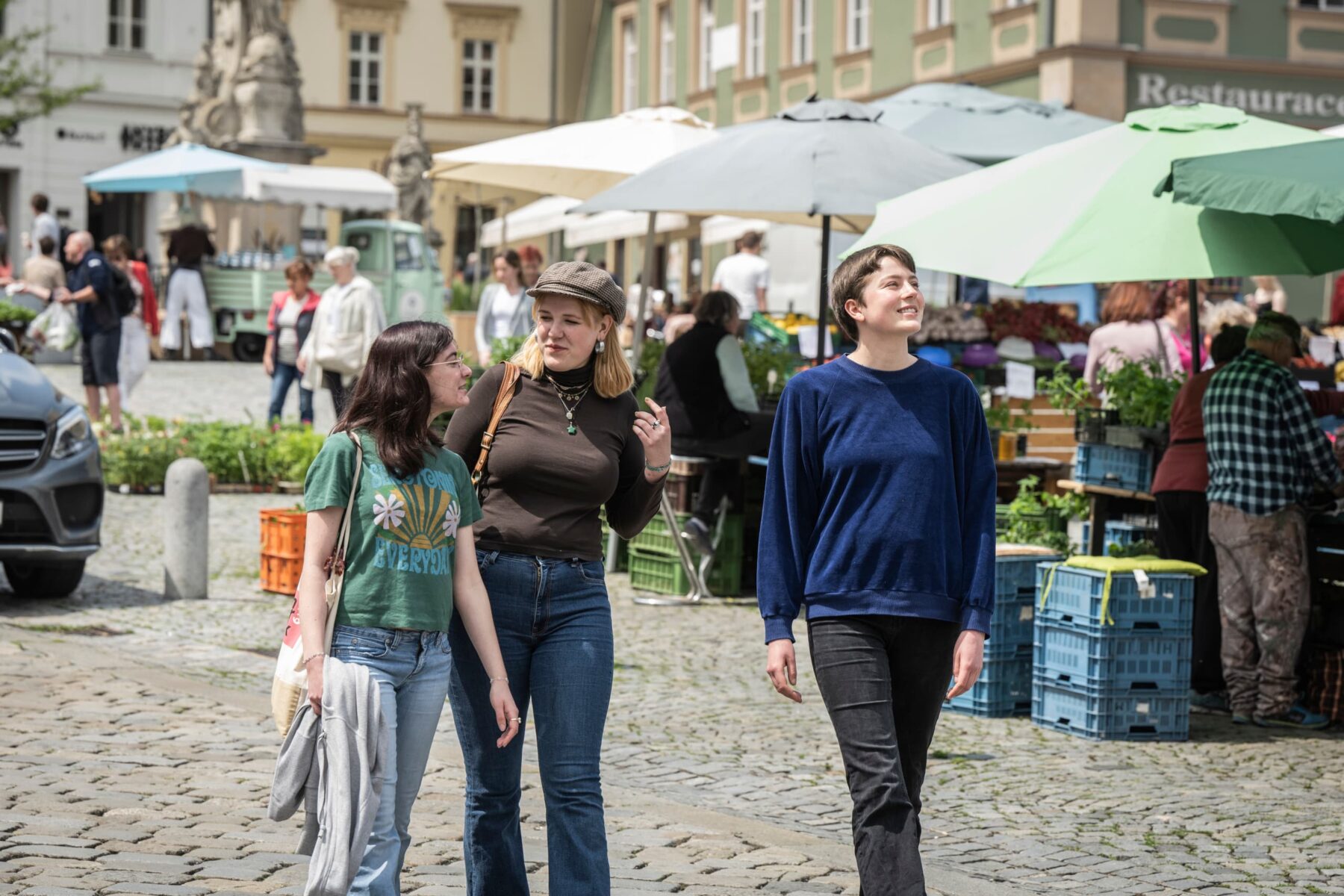 Drei junge Freiwillige schlendern lachend über einen Marktplatz in Brünn, bunte Marktstände und Sonnenschirme im Hintergrund.