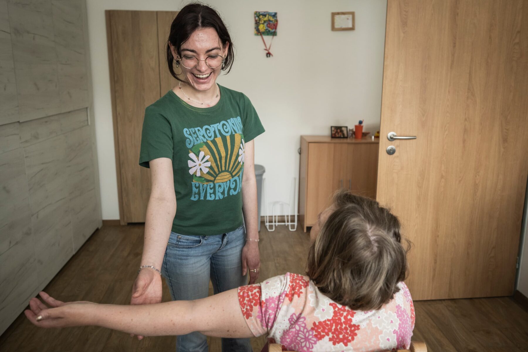 ASF‑Freiwillige in Brno: lächelnde junge Frau im grünen T‑Shirt reicht Hand zu einer sitzenden Frau im bunten Blumenoberteil