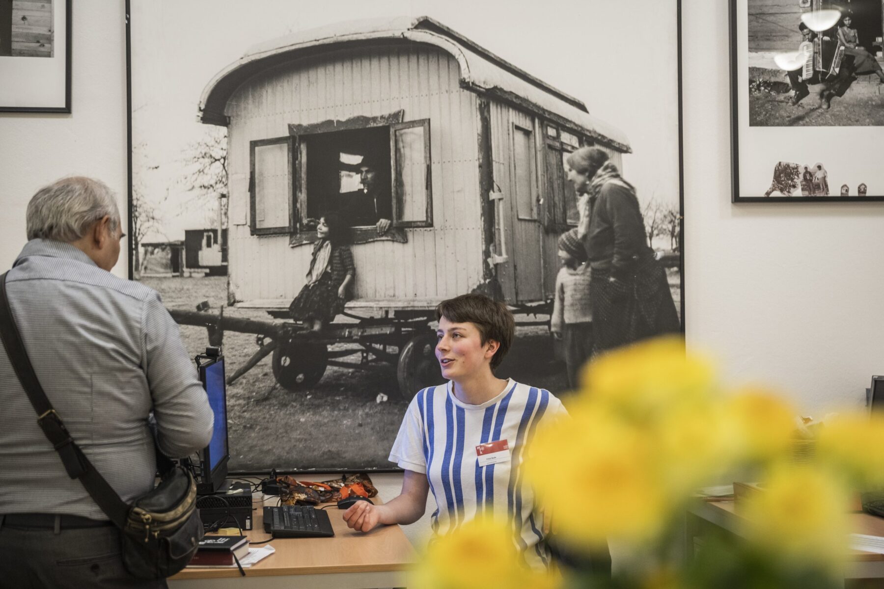 Junge Freiwillige am Tresen spricht mit älterem Mann; großes SW-Foto an der Wand, unscharfe gelbe Blumen im Vordergrund