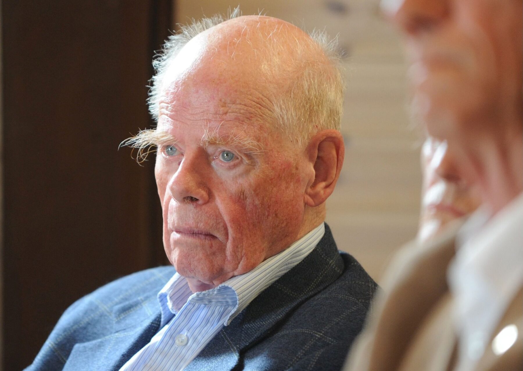 Älterer Mann mit buschigen weißen Augenbrauen und ernster Miene, blaues Sakko, gestreiftes Hemd, Seitenporträt.