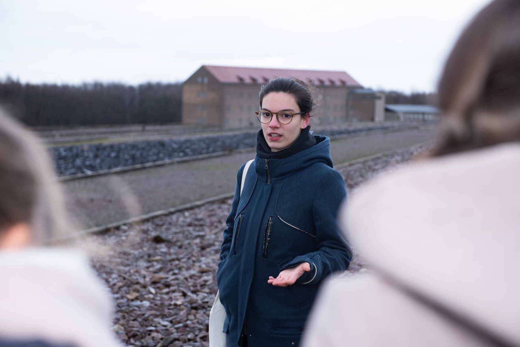 Junge Führerin mit Brille und dunklem Mantel spricht vor der Gedenkstätte Buchenwald, verschwommene Zuhörer im Vordergrund.