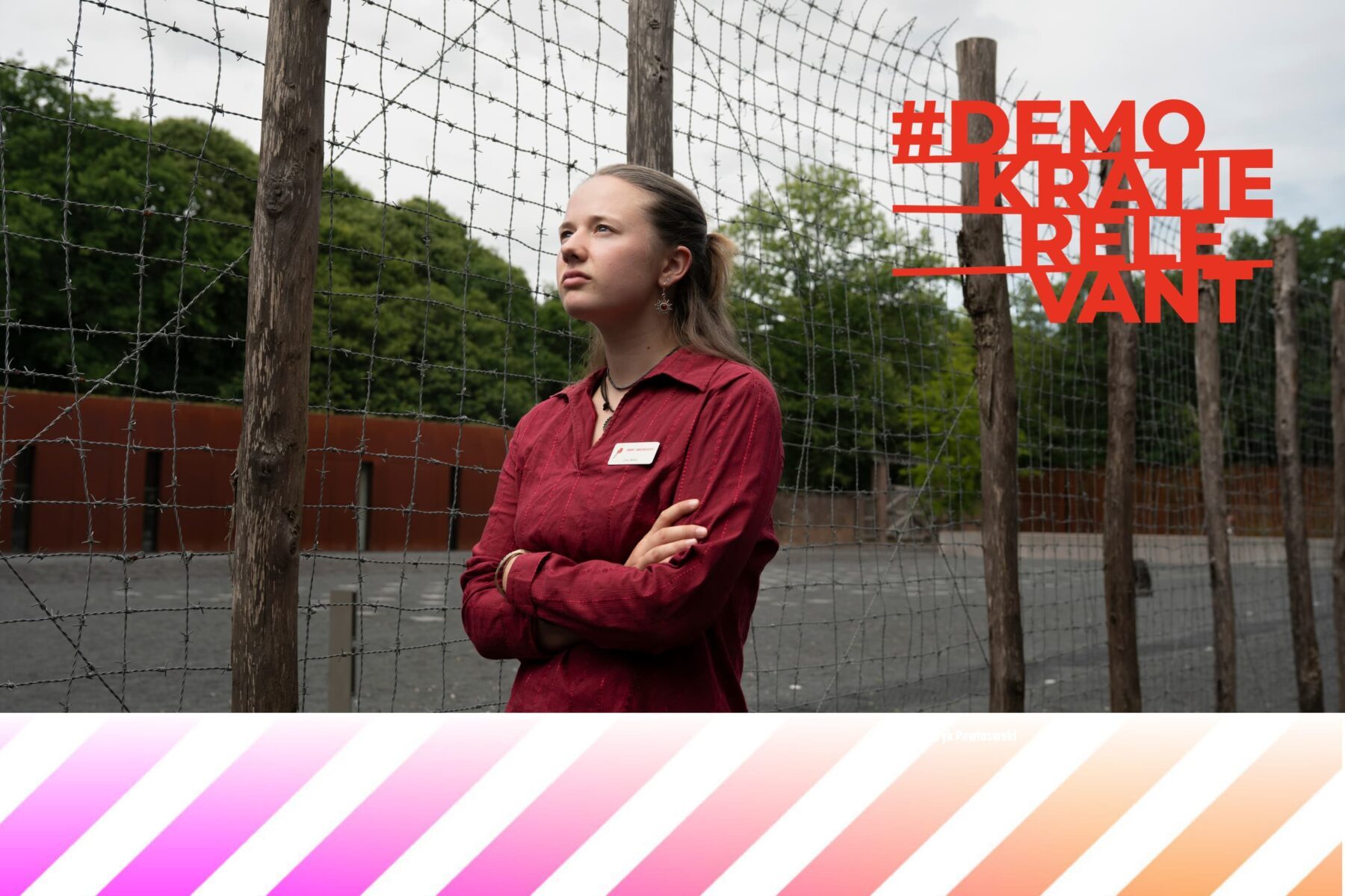 Frau in roter Bluse mit verschränkten Armen vor Stacheldrahtzaun, Blick aufwärts; #DEMOKRATIERELEVANT rechts oben.