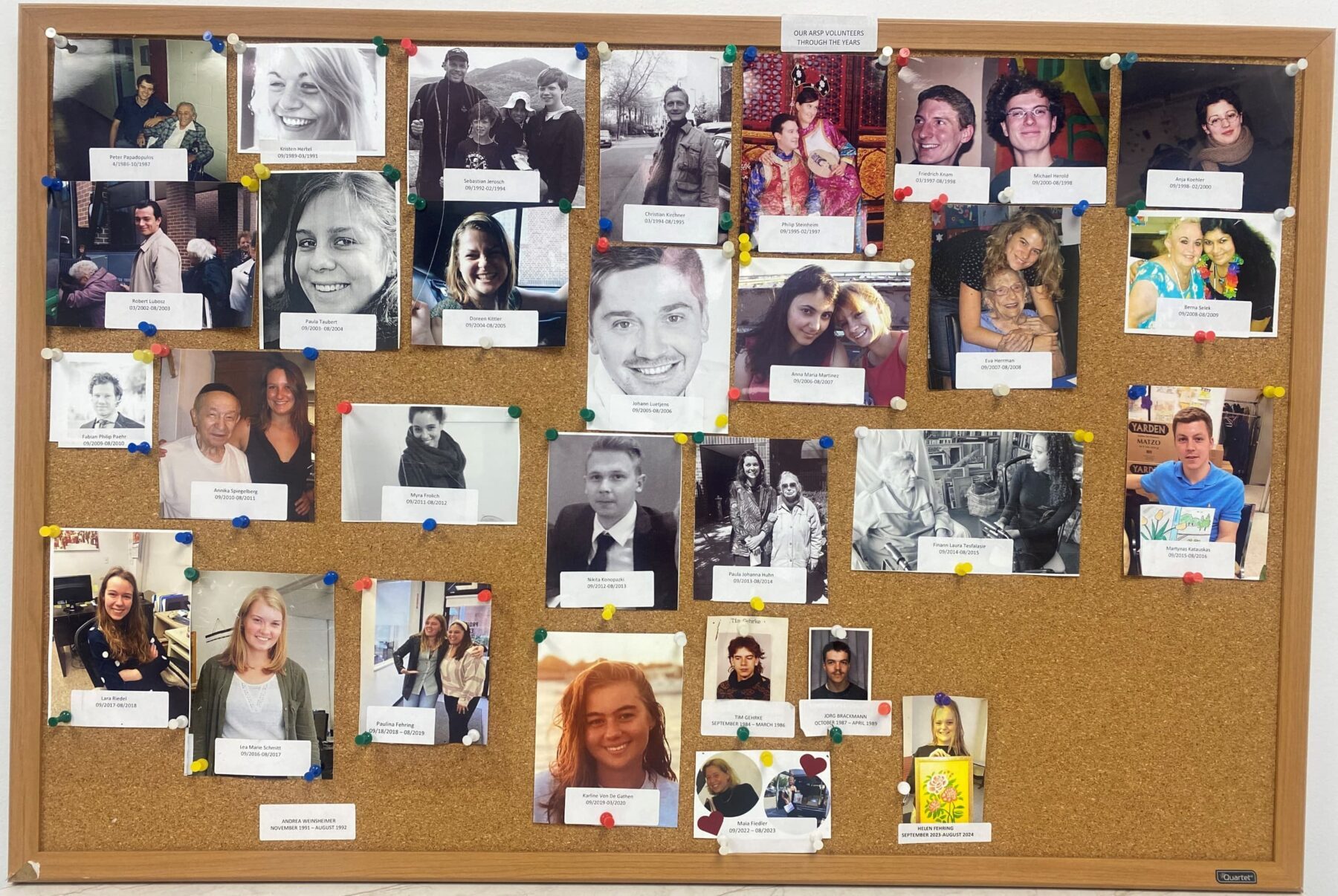 Kork-Pinnwand mit zahlreichen Porträtfotos und beschrifteten Karten, mit bunten Reißnägeln befestigt.