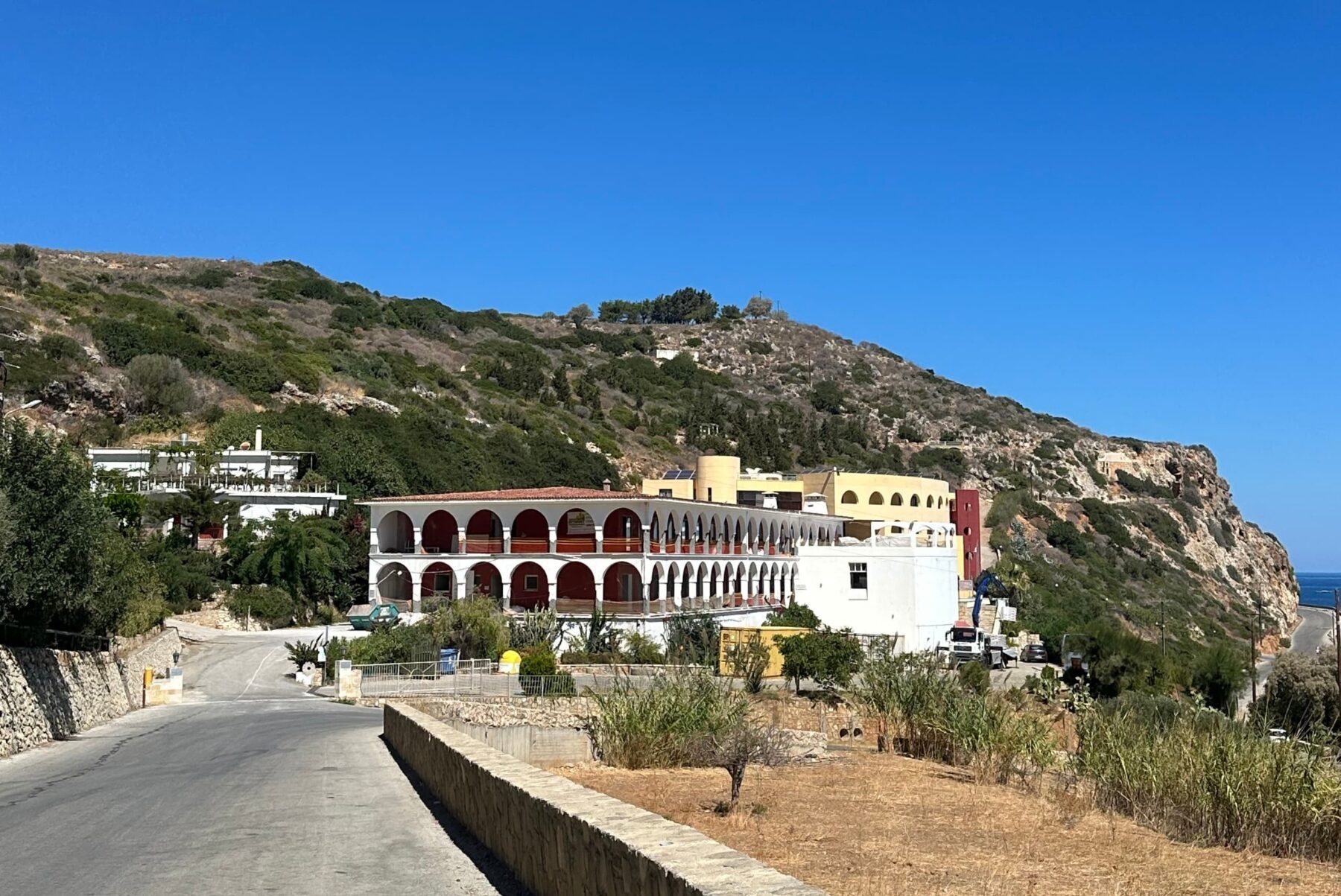 Mehrstöckiges Arkaden-Gebäude neben Küstenstraße, felsiger Hügel im Hintergrund, trockene Vegetation und klarer blauer Himmel.