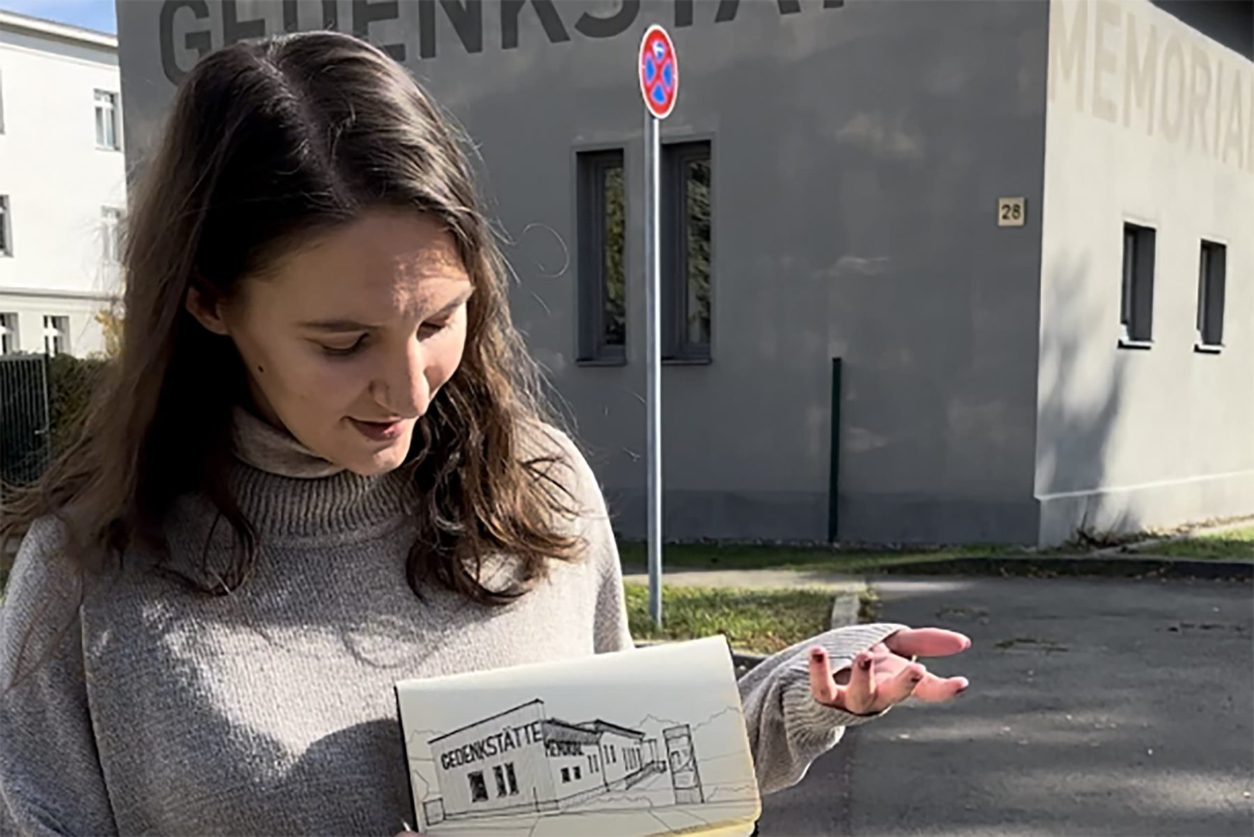 Женщина держит блокнот с рисунком мемориального комплекса. На заднем плане здание с надписью GEDENKSTÄTTE.