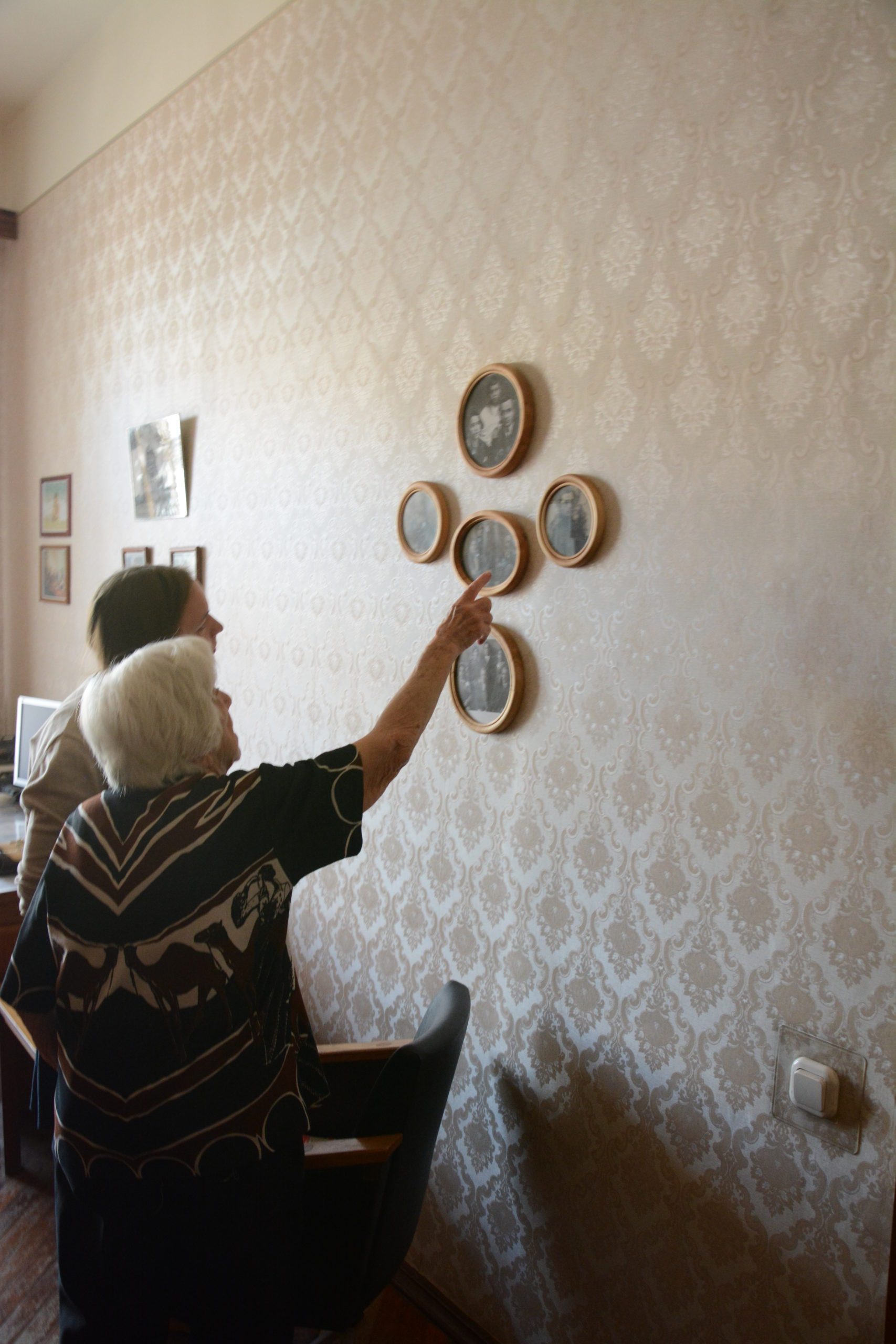 Две женщины смотрят на настенные фотографии в круглых рамках в комнате с узорчатыми обоями.
