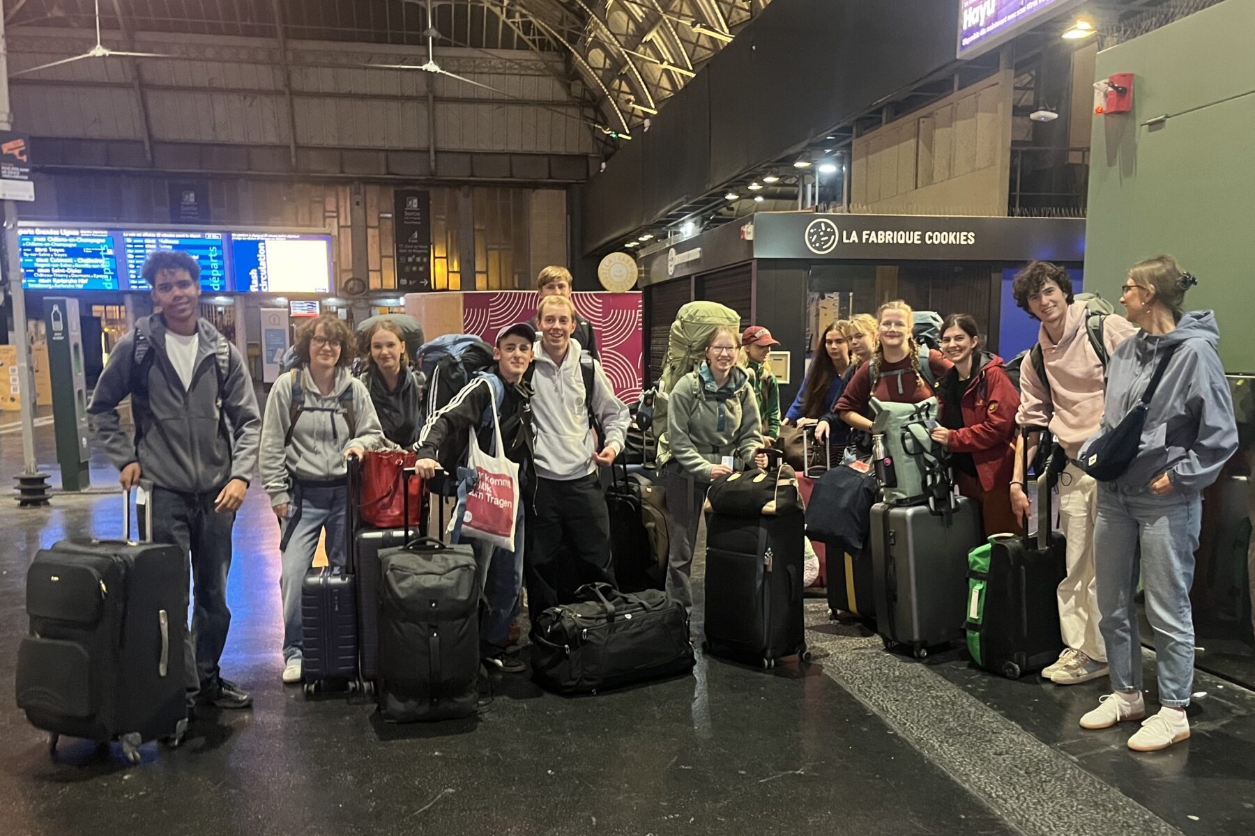 Groupe d'une douzaine de jeunes voyageurs souriants en gare, entourés de valises et sacs devant la boutique 'La Fabrique Cookies'.