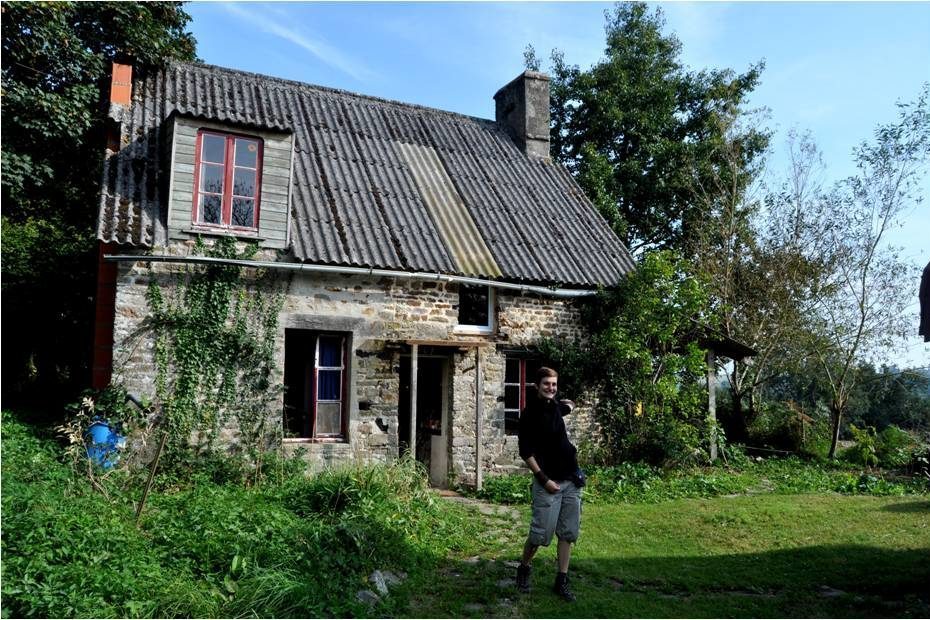 Bénévole ASF souriant devant une maison en pierre à Saint-Jean-le-Blanc, toit ondulé, lierre et jardin.