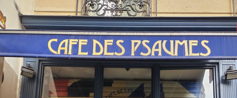 Serveuse souriante tenant un plateau avec tasses rouges et vertes dans l'embrasure du Café des Psaumes, terrasse animée.