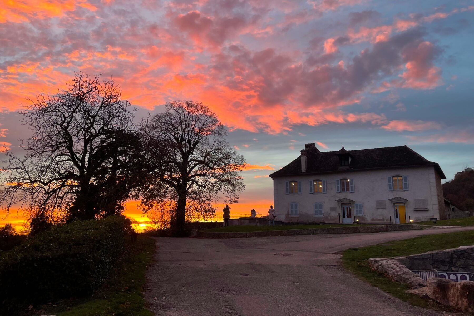 Maison d'Izieu au coucher du soleil, ciel rose-orangé, arbres nus en silhouette et visiteurs sur la terrasse.