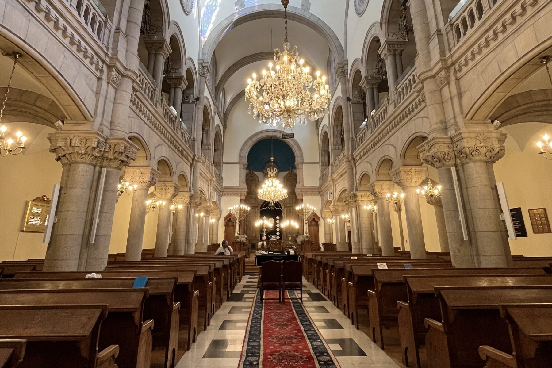 Intérieur de la Grande Synagogue de Lyon: nef centrale avec tapis rouge, bancs en bois, colonnes en pierre et grands lustres.