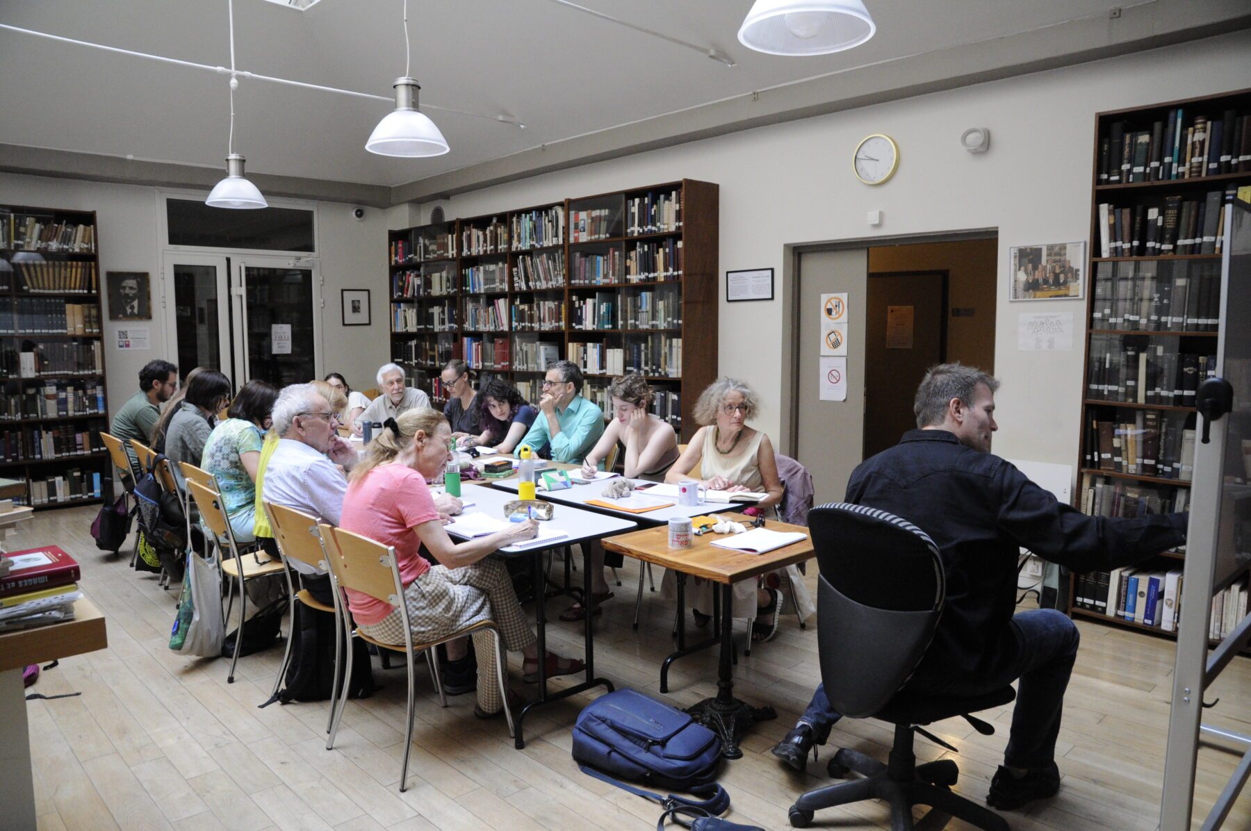 Groupe d'adultes en cours asf MCY dans une bibliothèque parisienne, assis autour de tables, prof à droite devant tableau.