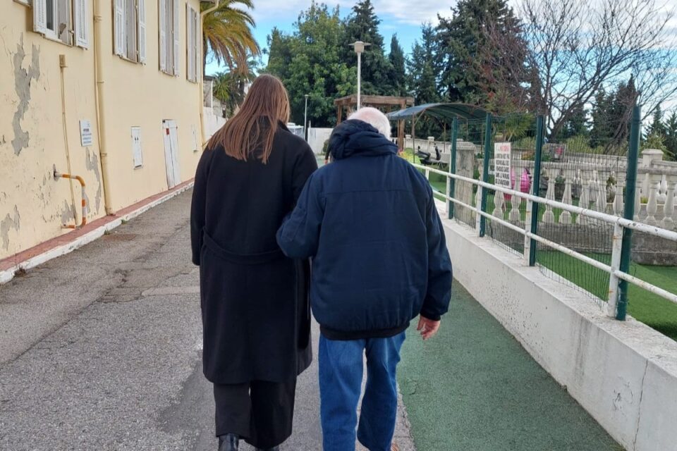 Femme et homme âgé marchant bras liés le long d'une allée à La Colline (Nice), mur jaune à gauche, clôture verte à droite.
