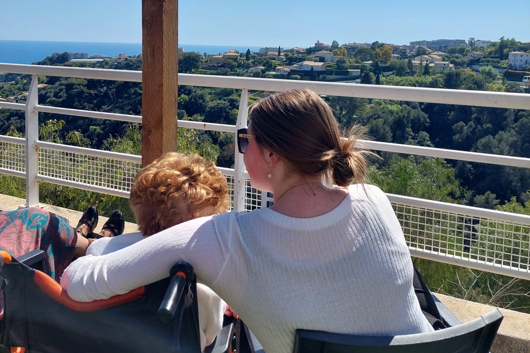 Jeune femme embrassant une personne âgée en fauteuil sur une terrasse, vue sur la colline et la mer de Nice.