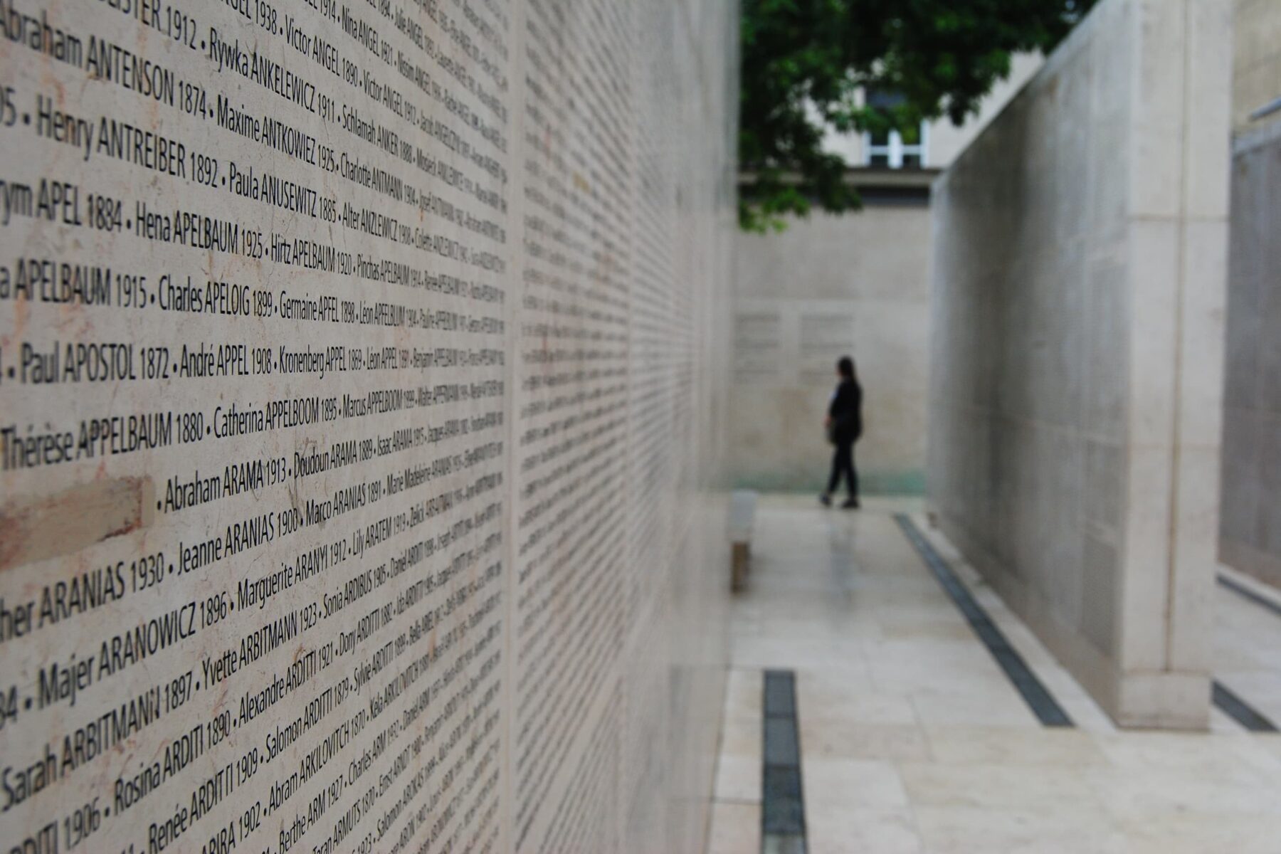 Mur du Mémorial de la Shoah à Paris gravé de noms, couloir en pierre et personne seule floue au loin