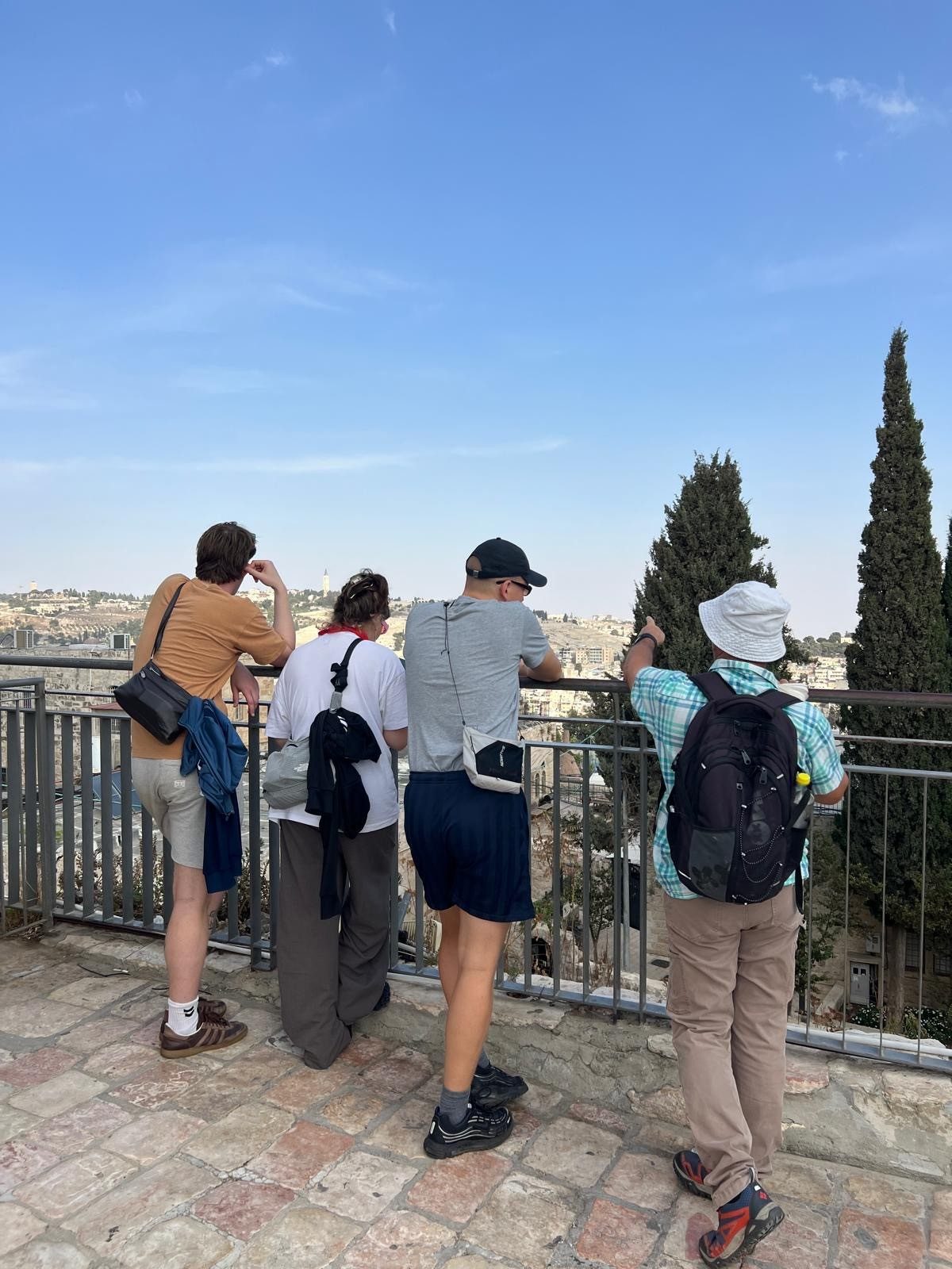 Four people leaning on a railing, overlooking a sunlit city from a stone terrace with cypress trees under a clear blue sky.