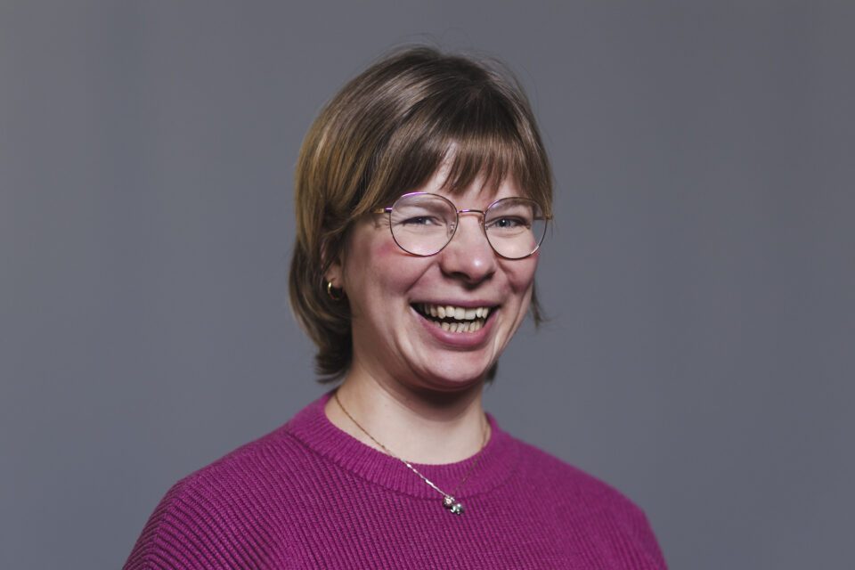 Kvinne i rosa genser med runde briller og kort brunt hår smiler bredt foran nøytral grå bakgrunn.